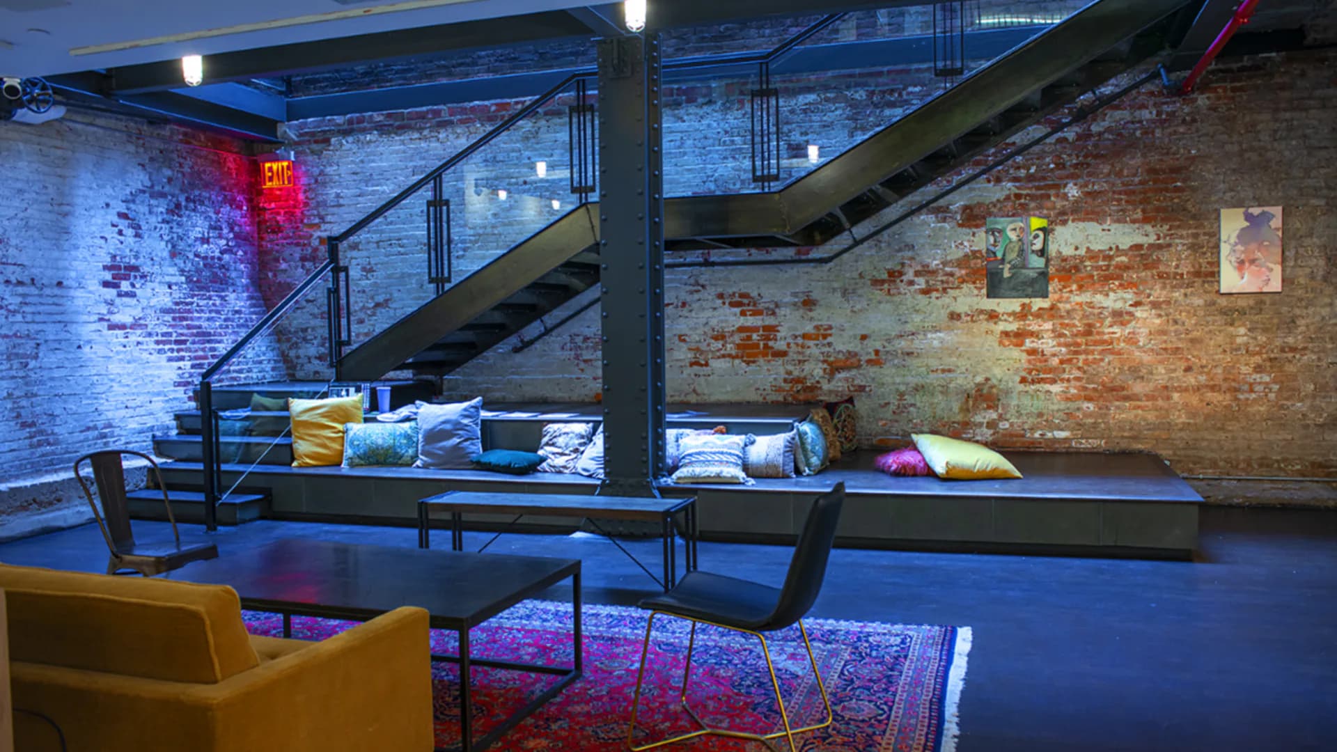 Modern gallery interior at Pen + Brush with exposed brick, industrial ceiling, blue lighting, colorful cushions, and artworks on walls.