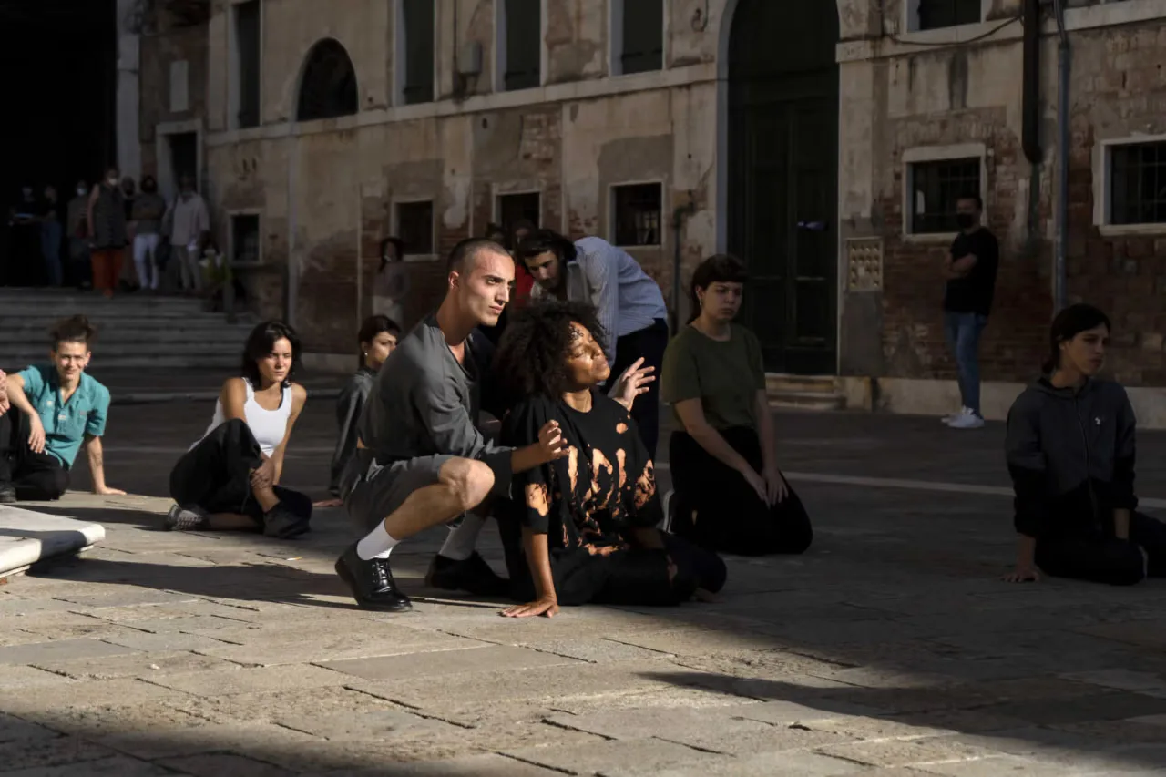 Performance or installation in historic plaza with seated and standing participants in dark clothing, natural daylight, stone architecture background.