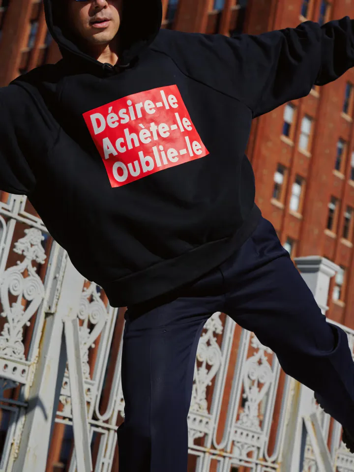 Person in black hoodie with red text box graphic stretched against brick building, arm raised in dynamic pose.