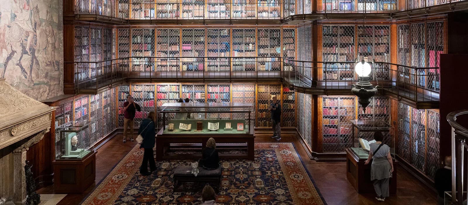 Interior of a historic library room with floor-to-ceiling wooden bookcases filled with leather-bound volumes, central display case, ornate rug, and period furniture.