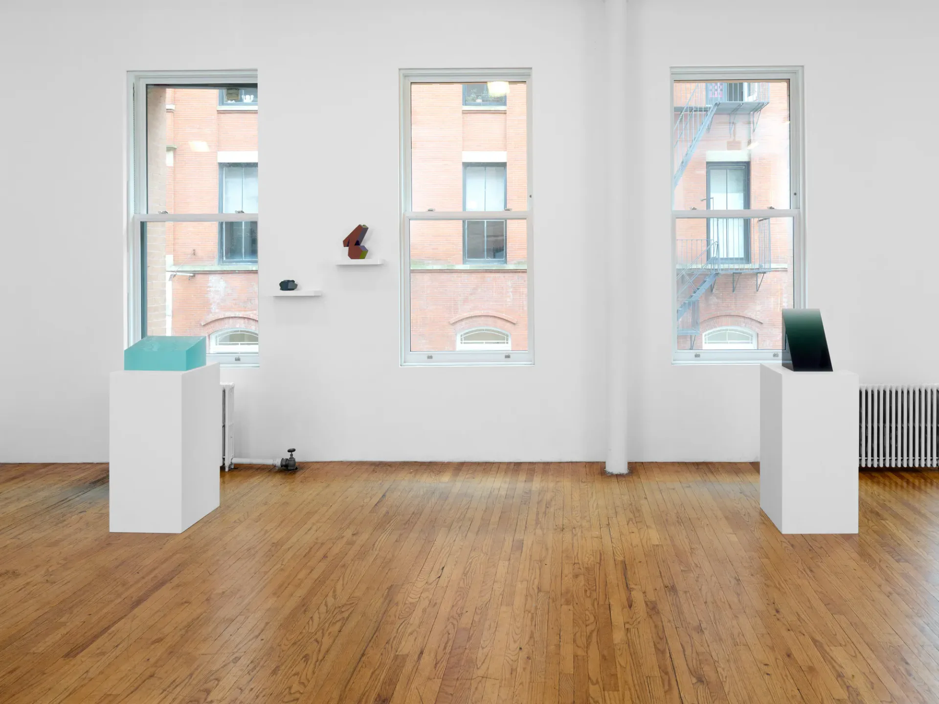 Bright gallery interior with wooden floor, three large windows overlooking pink building, white partition walls with mounted artworks including turquoise sculptural form, minimal furniture.