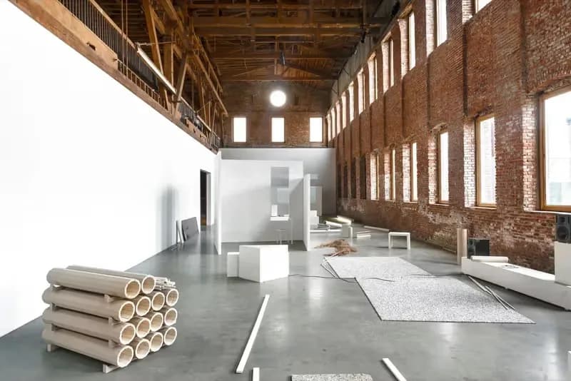 Industrial brick warehouse interior with exposed wooden ceiling beams, large windows, and construction materials including rolled tubes and sheets on the concrete floor.
