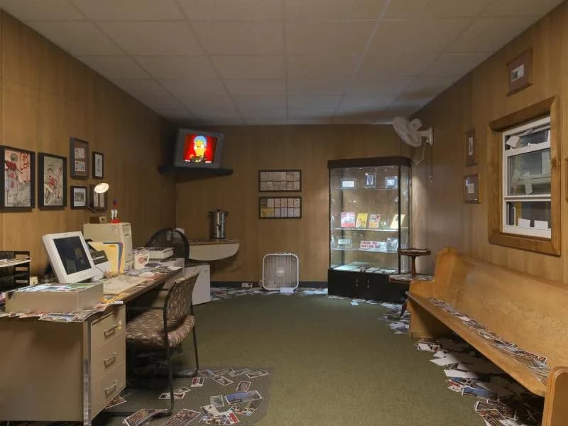 Museum or gallery office space with tan walls, desk with computer, display cases, glowing red neon sign, and scattered papers on floor.
