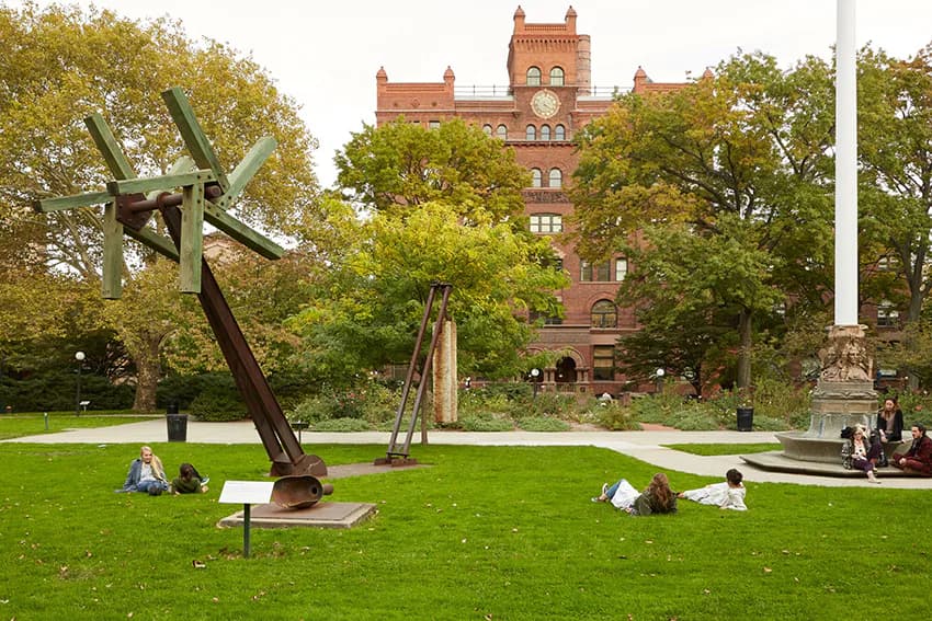 Pratt Institute Sculpture Park grounds featuring dark metal abstract sculptures on grass with Gothic Revival brick tower and mature trees.