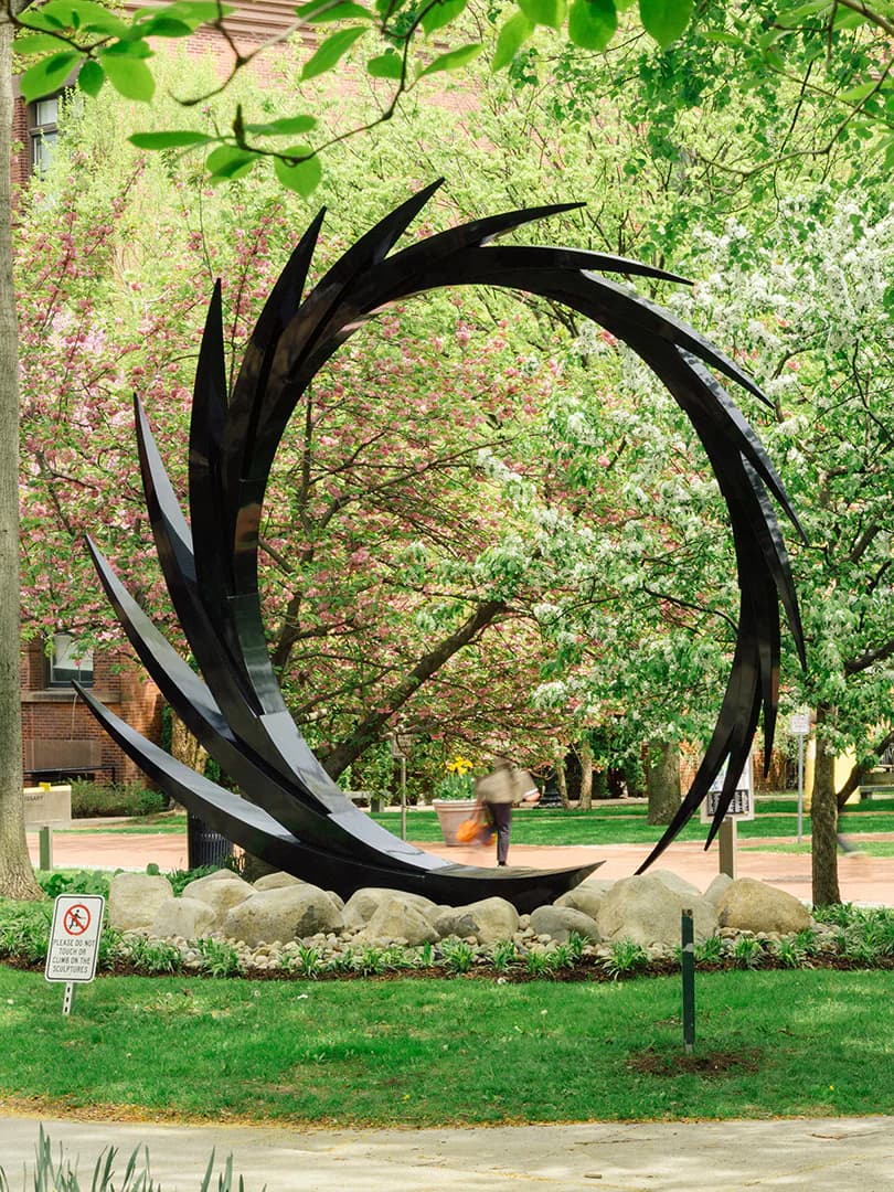Large black metal abstract circular sculpture with sweeping curved forms creating negative space; installed at Pratt Institute Sculpture Park.