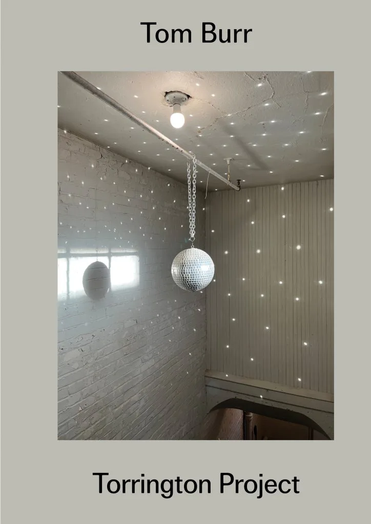 Interior installation by Tom Burr titled Torrington Project featuring a suspended mirrored disco ball with chain in a sparse room with recessed lighting and vertical blinds.