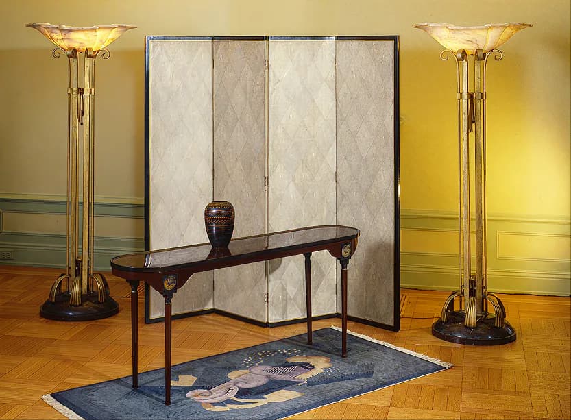 Gallery interior with yellow walls, wooden floor, tall brass floor lamps, decorative room divider, dark wooden console table, and blue patterned rug.