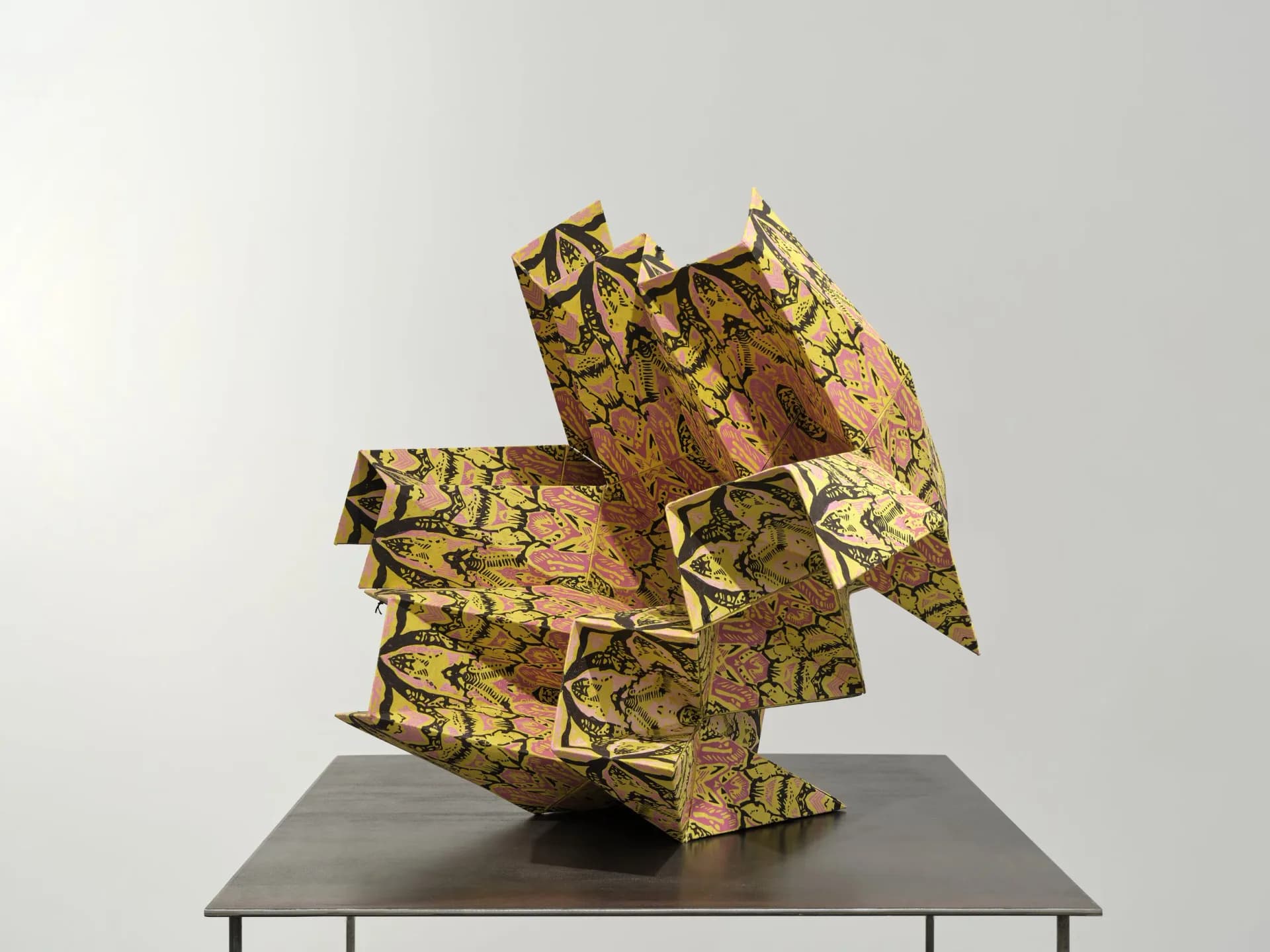 Abstract sculptural form folded from yellow-green and rust patterned fabric or paper, positioned on gray platform against neutral background.