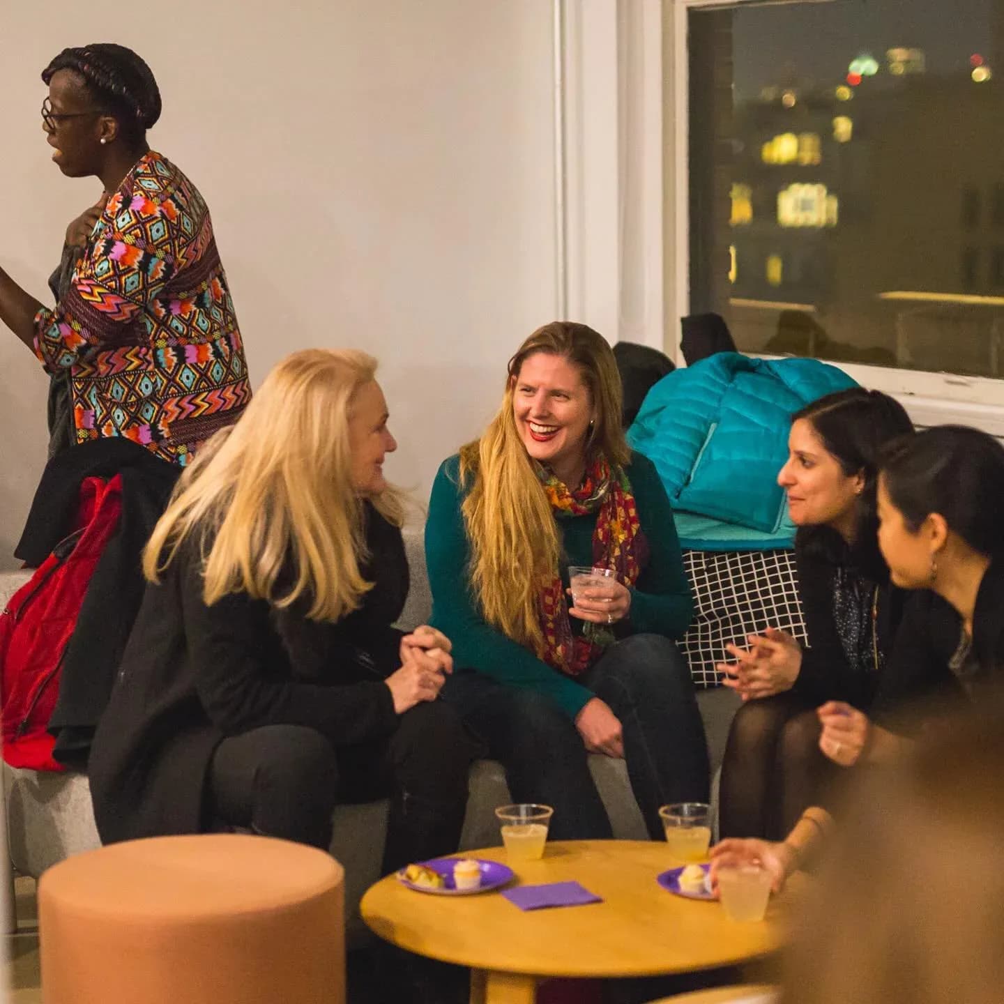 People socializing indoors at an event with a yellow side table, drinks, and colorful furnishings visible.