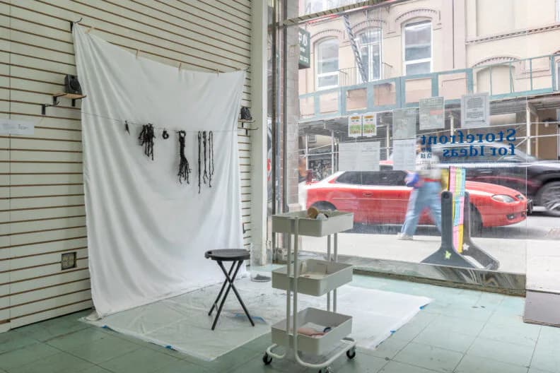 Gallery space with white hanging fabric, clothing items pinned to fabric backdrop, metal stool, wooden frame structure, and city street with red car visible outside.