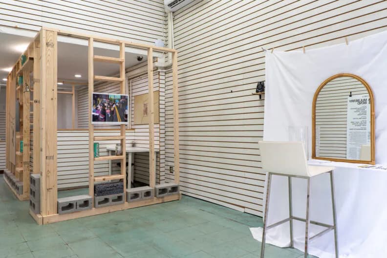 Gallery interior with corrugated white walls, large wooden grid-frame display cabinet with glass panels containing photographs, arched mirror, and metal stool.