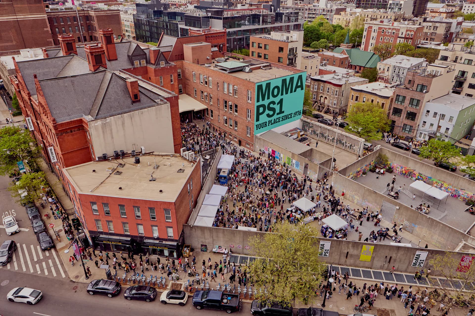 Aerial view of P.S.1 Contemporary Art Center's plaza in Queens with large crowds gathered, red brick building with green MoMA PS1 signage visible.