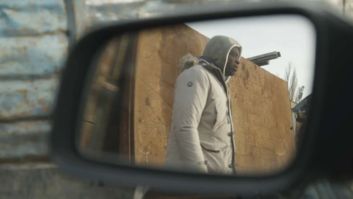 Figure in light-colored clothing photographed through a vehicle's side mirror against a tan/brown wall.
