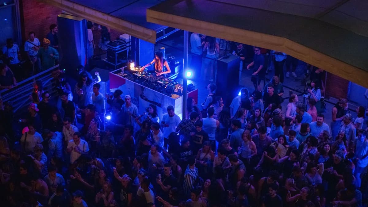 Overhead view of gallery interior with crowd gathered on multi-level floor under blue stage lighting with performance setup.