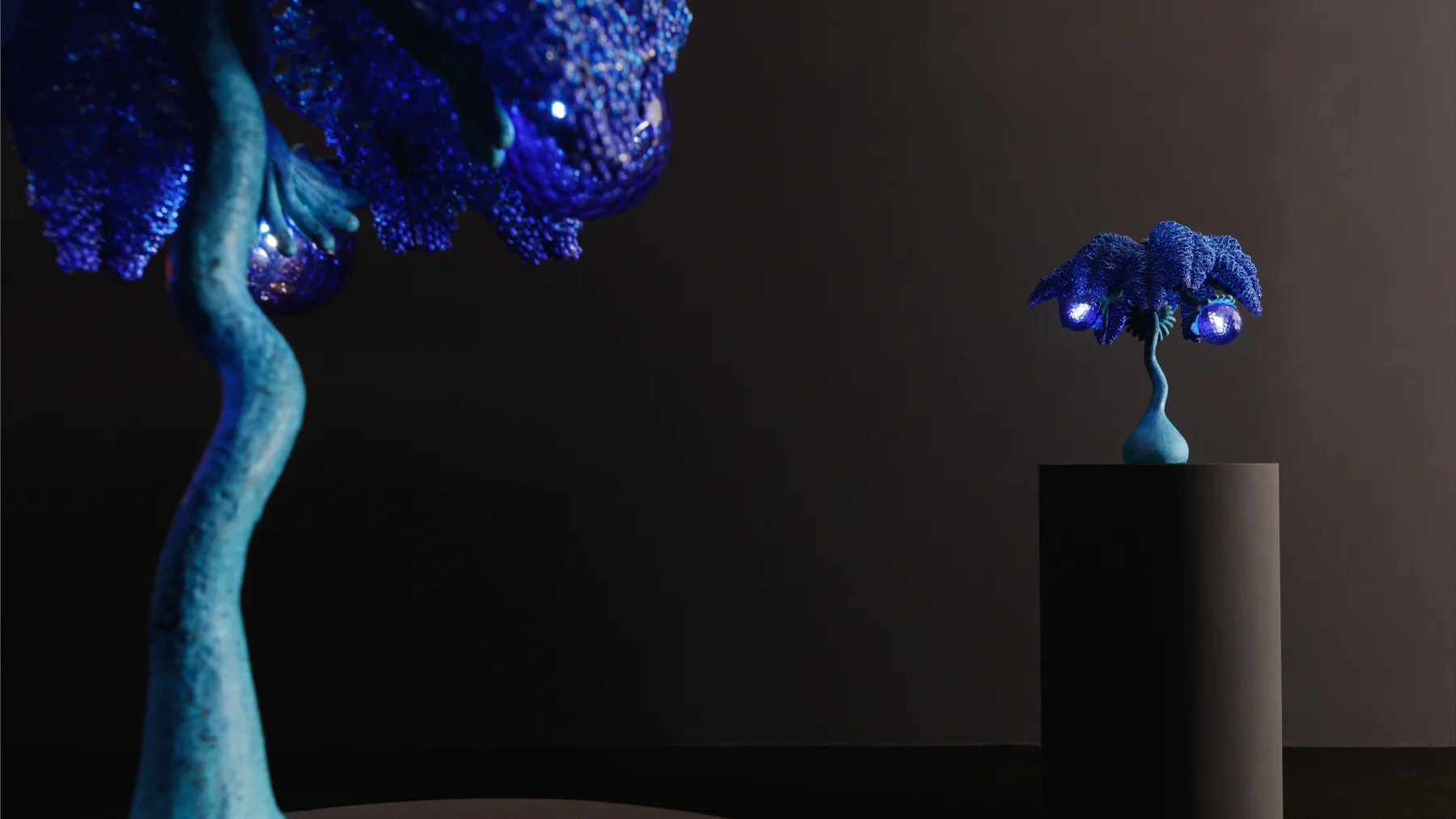 Two sculptural trees with luminescent blue crystalline foliage and pale turquoise trunks against a dark background, one large on left and one small on right pedestal.