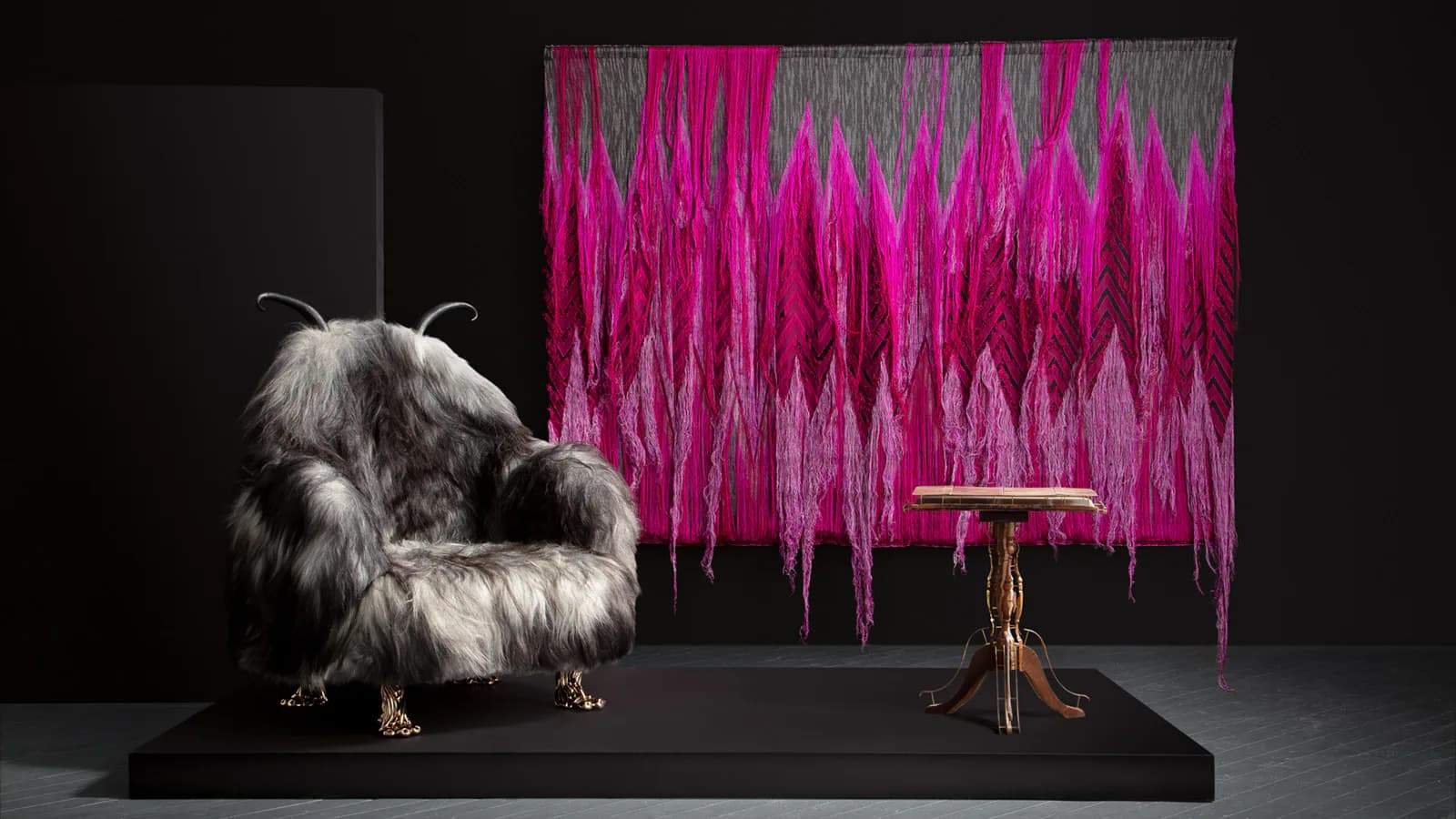Gallery installation featuring a white furry sculptural creature on black platform beside a wooden chair facing a vibrant magenta sound-wave textile wall.