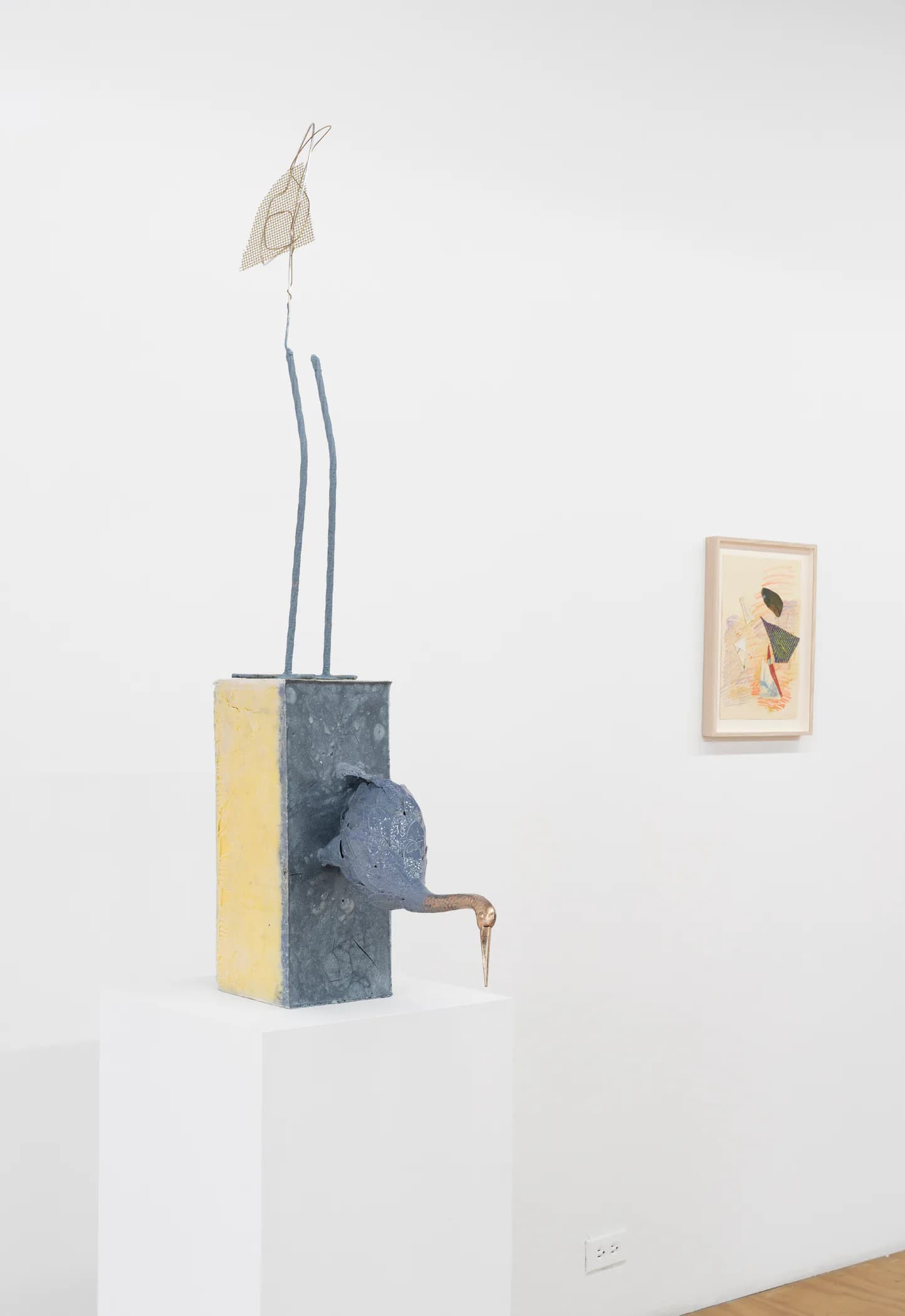 Mixed-media sculpture on white pedestal featuring blue and yellow geometric base with protruding elements, suspended strings with leaf form above, and small framed drawing on adjacent wall.
