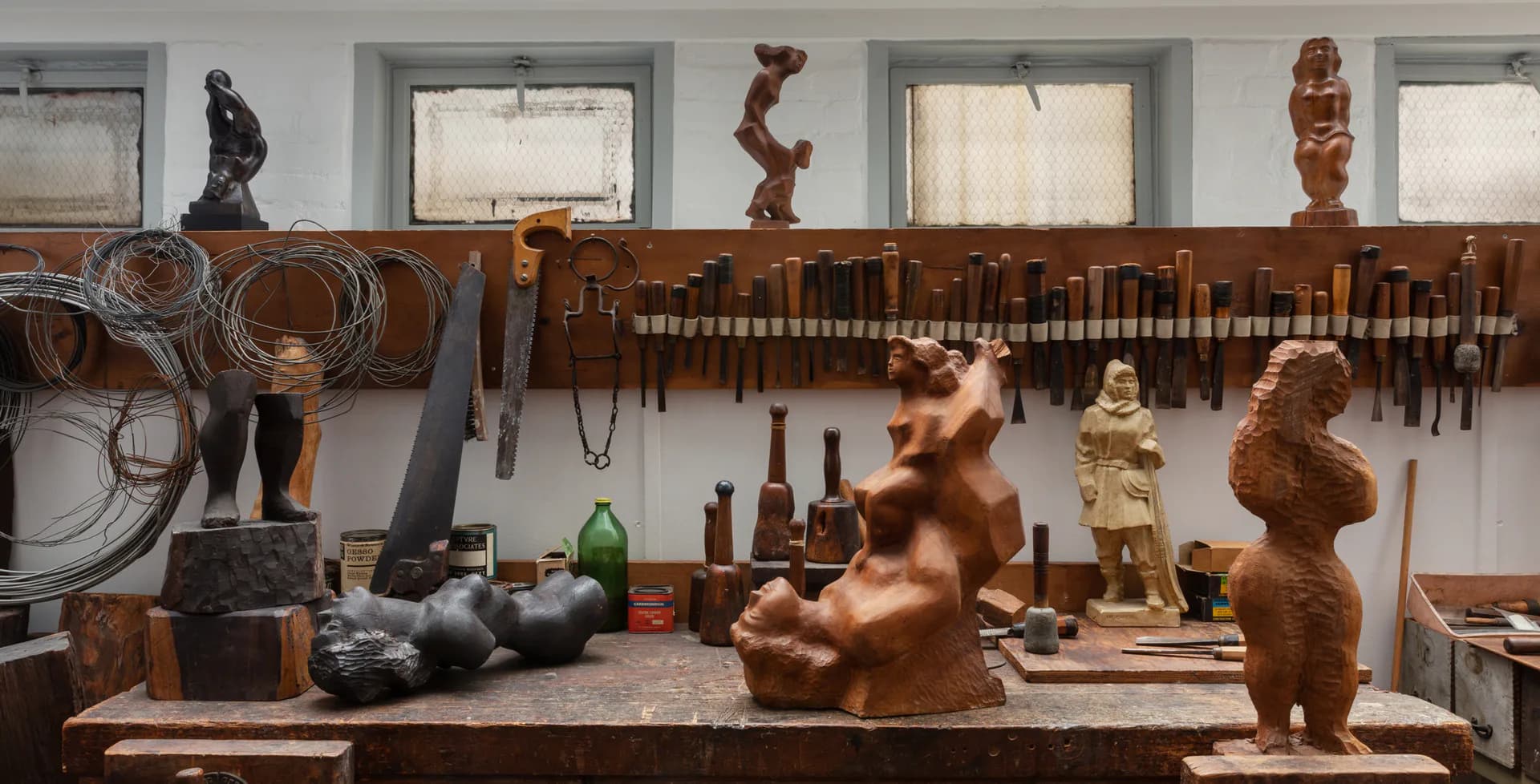 Sculptor's studio workbench displaying wooden figurative sculptures in progress, carving tools, and reference materials against white walls with organized tool storage.