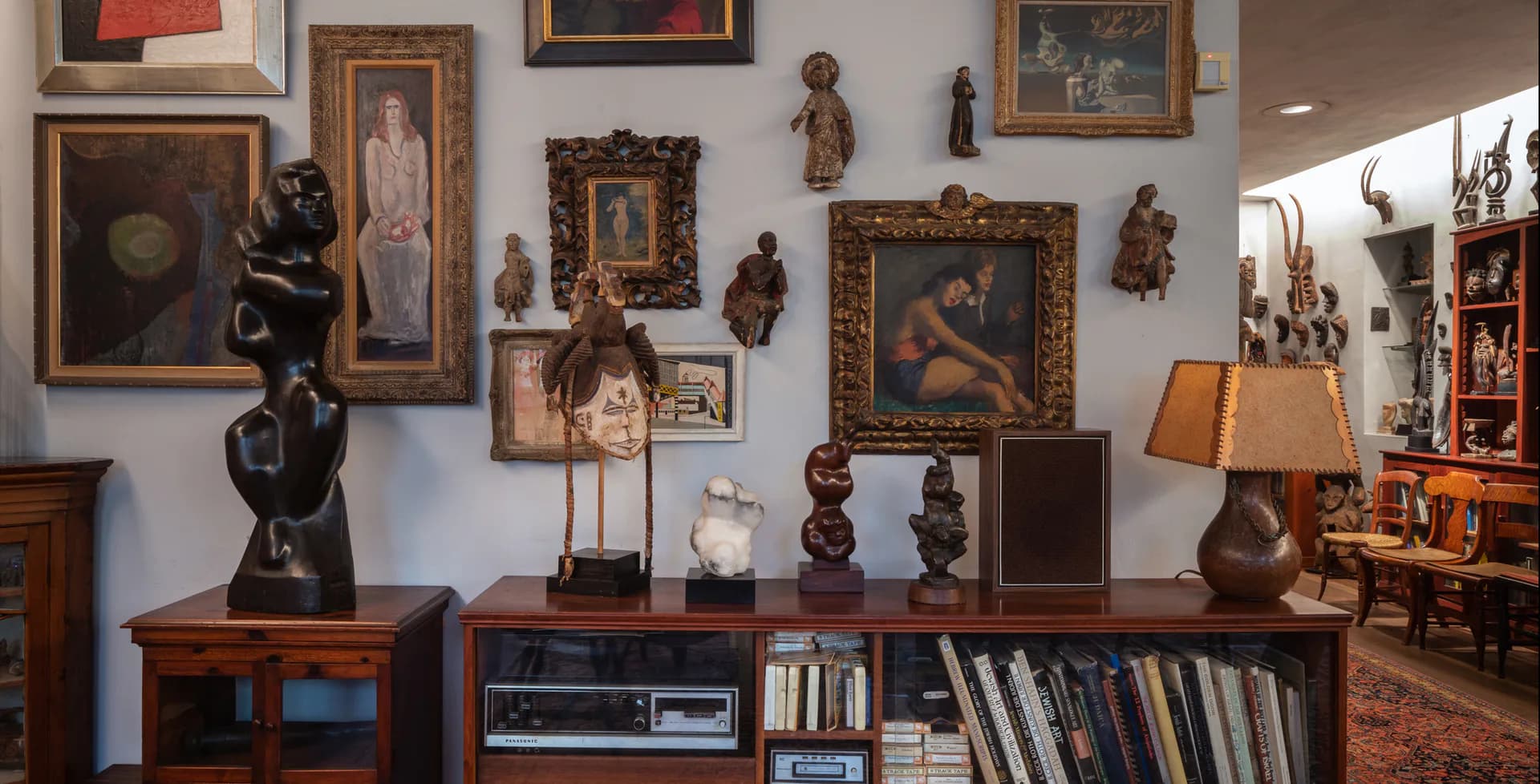 Gallery room with framed paintings and sculptures displayed on white walls, wooden furniture pieces, bronze figurative works, and books on lower shelving.