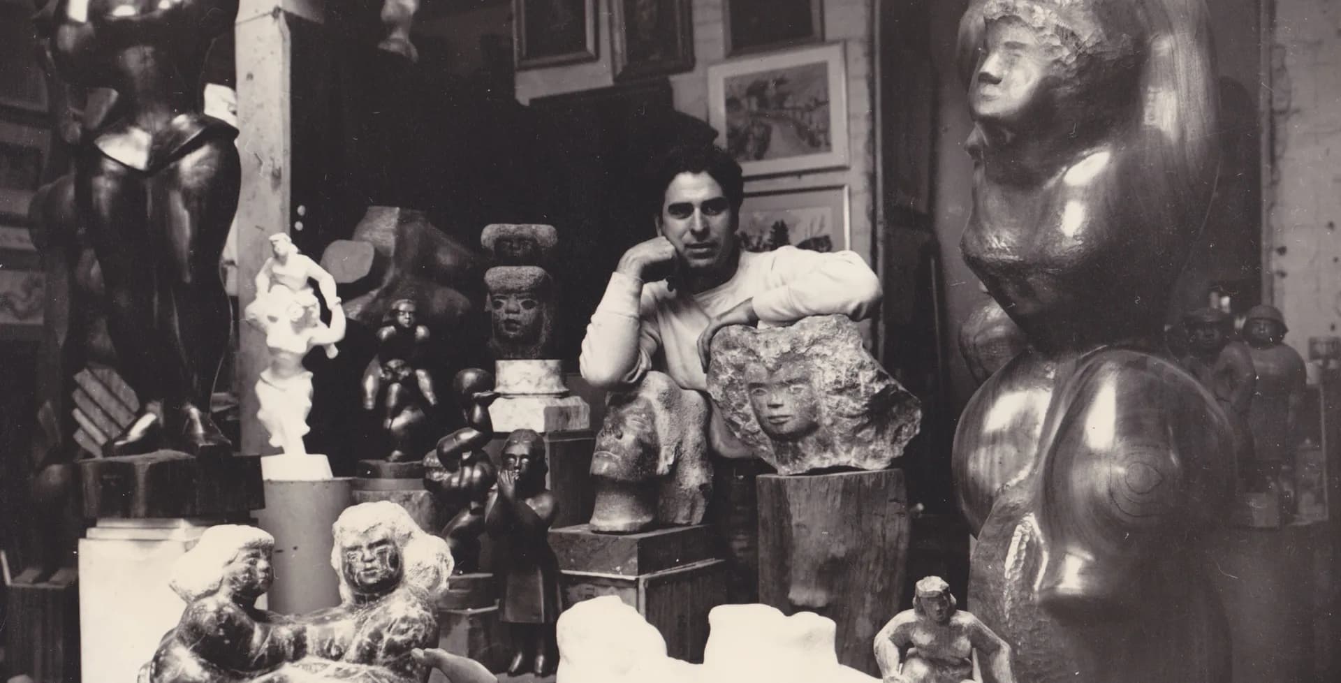 Black-and-white photograph of a studio interior with a seated sculptor surrounded by wooden and stone sculptures, busts, and art objects on shelves and surfaces.