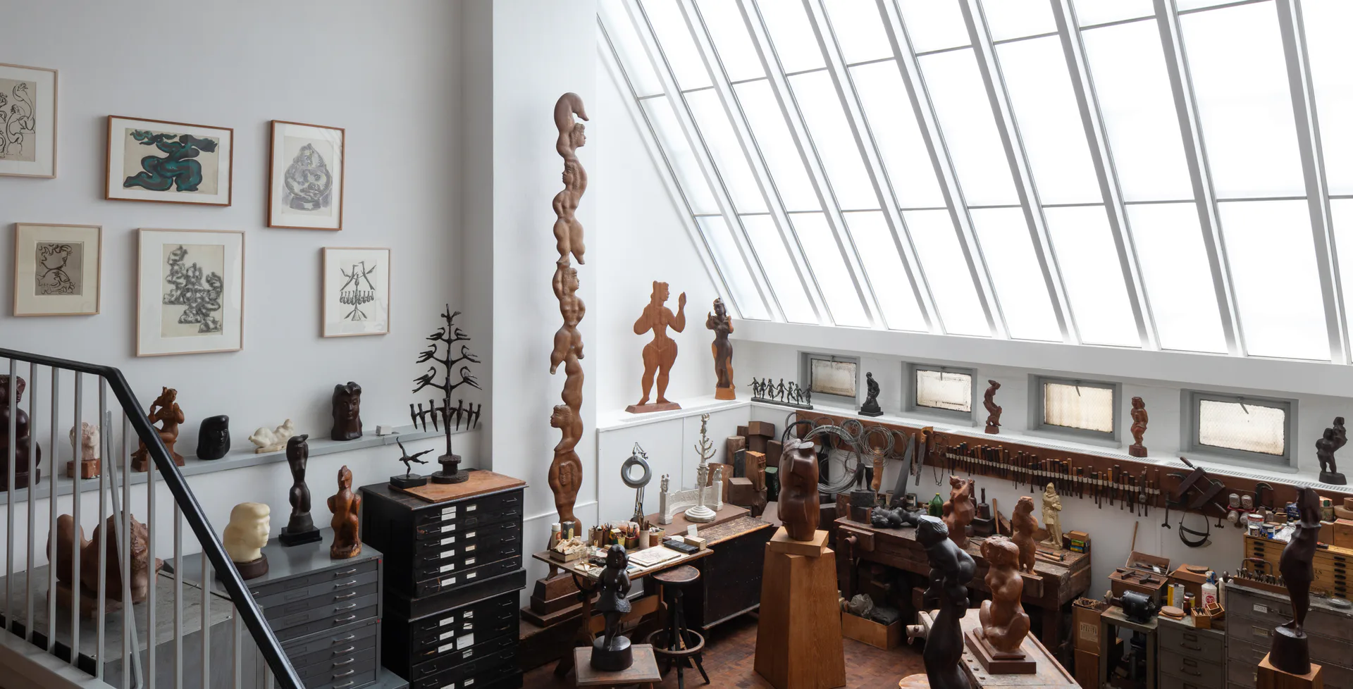 Bright modern gallery interior with skylights, wooden sculptures displayed on pedestals and shelves, framed drawings on walls, and metal railing along the upper level.