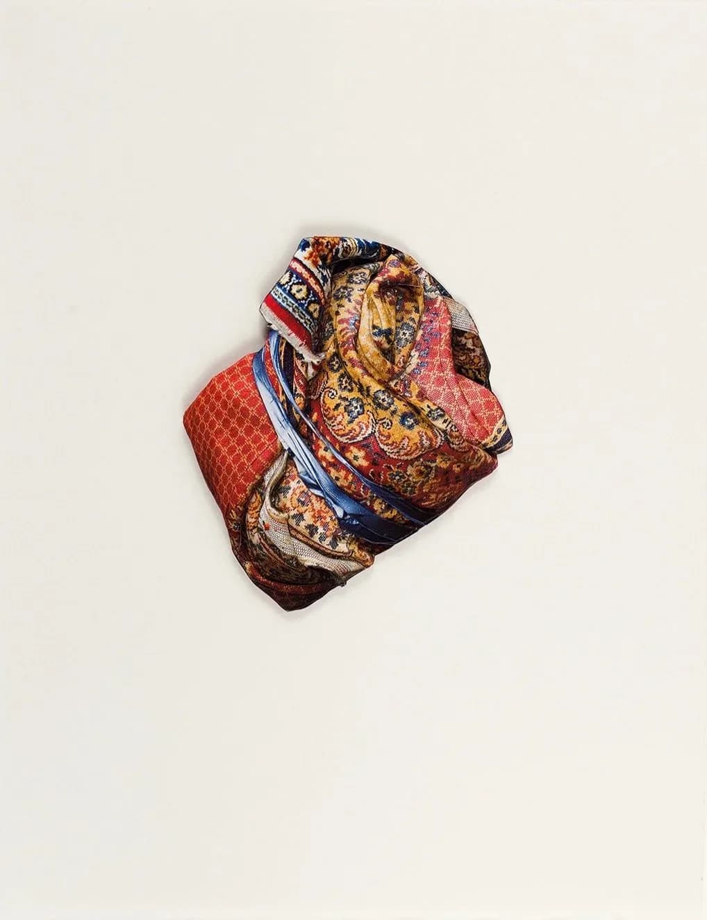 Crumpled or folded textile artwork in rich reds, golds, and blues with ornate patterning against white wall background.