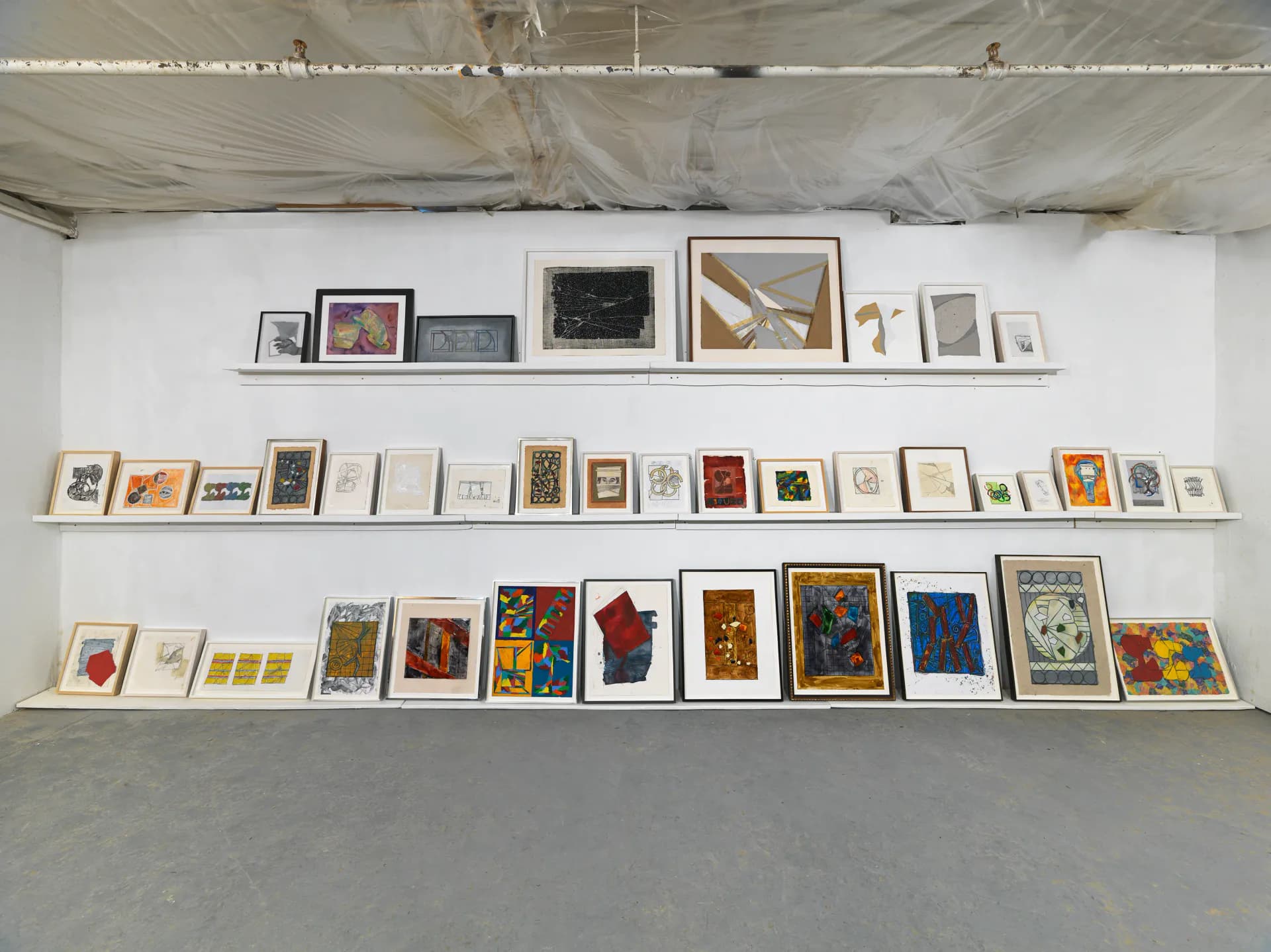 Gallery wall displaying three horizontal shelves lined with framed artworks and prints of varying sizes, including paintings, drawings, and mixed-media pieces in diverse styles and colors.