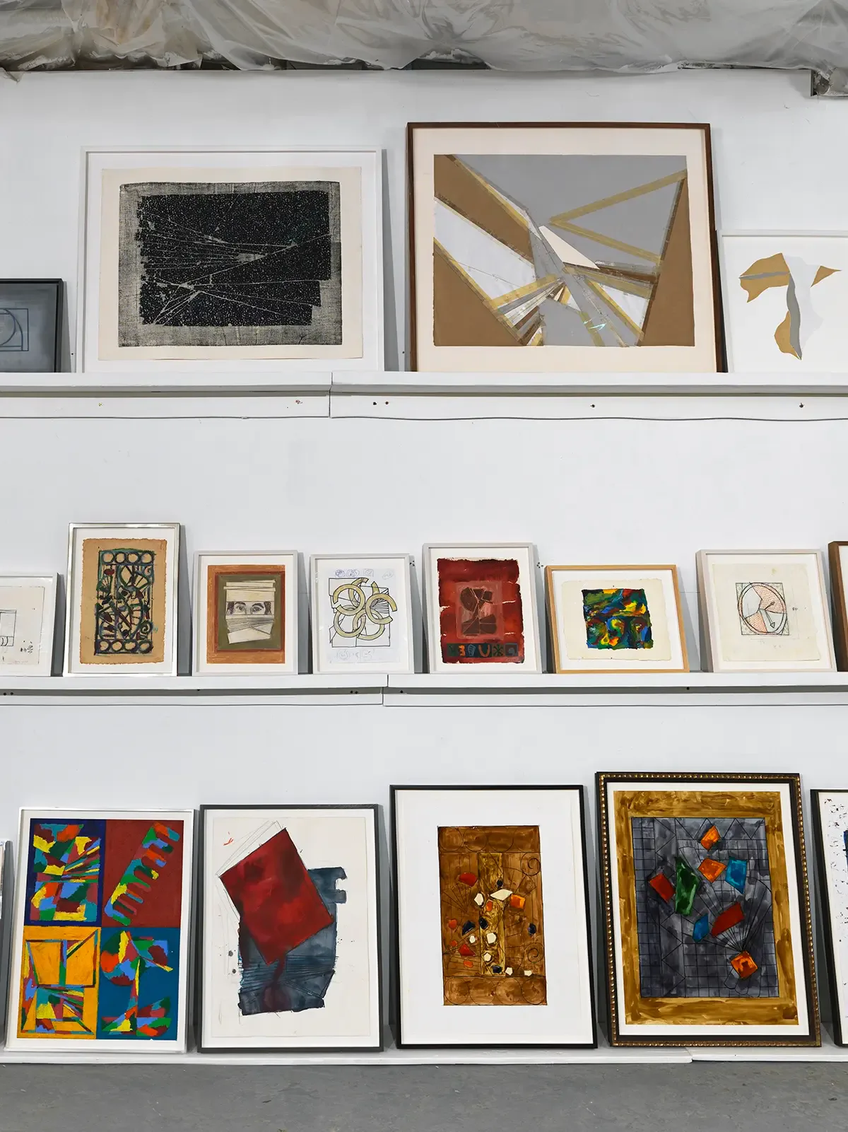 Gallery wall with three white shelves displaying framed artworks: top shelf features two large abstract pieces, middle and lower shelves show smaller framed paintings and prints in varied media.