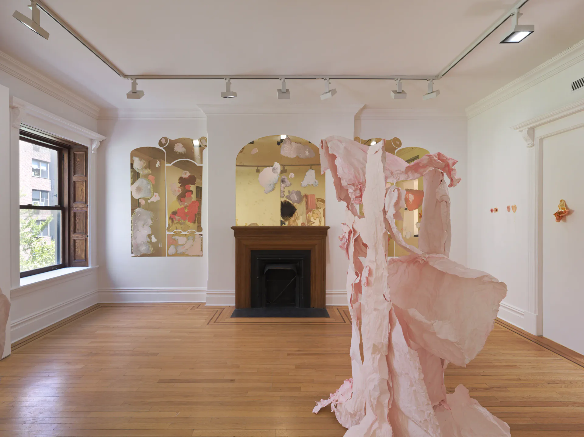 Gallery space with wooden fireplace, two large abstract mixed-media panel artworks on left wall, and pink fabric textile installation suspended in center of room.
