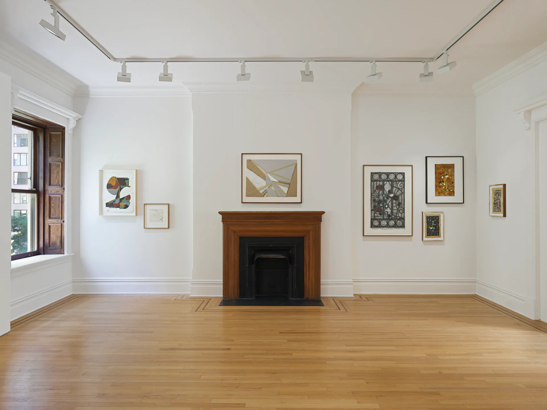 Gallery interior featuring wooden fireplace and various framed artworks hung on white walls: abstract drawings, prints, and paintings in assorted sizes and media.