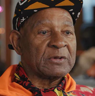 Portrait photograph of an older man wearing a patterned black and gold head wrap and orange shirt, looking directly at camera.