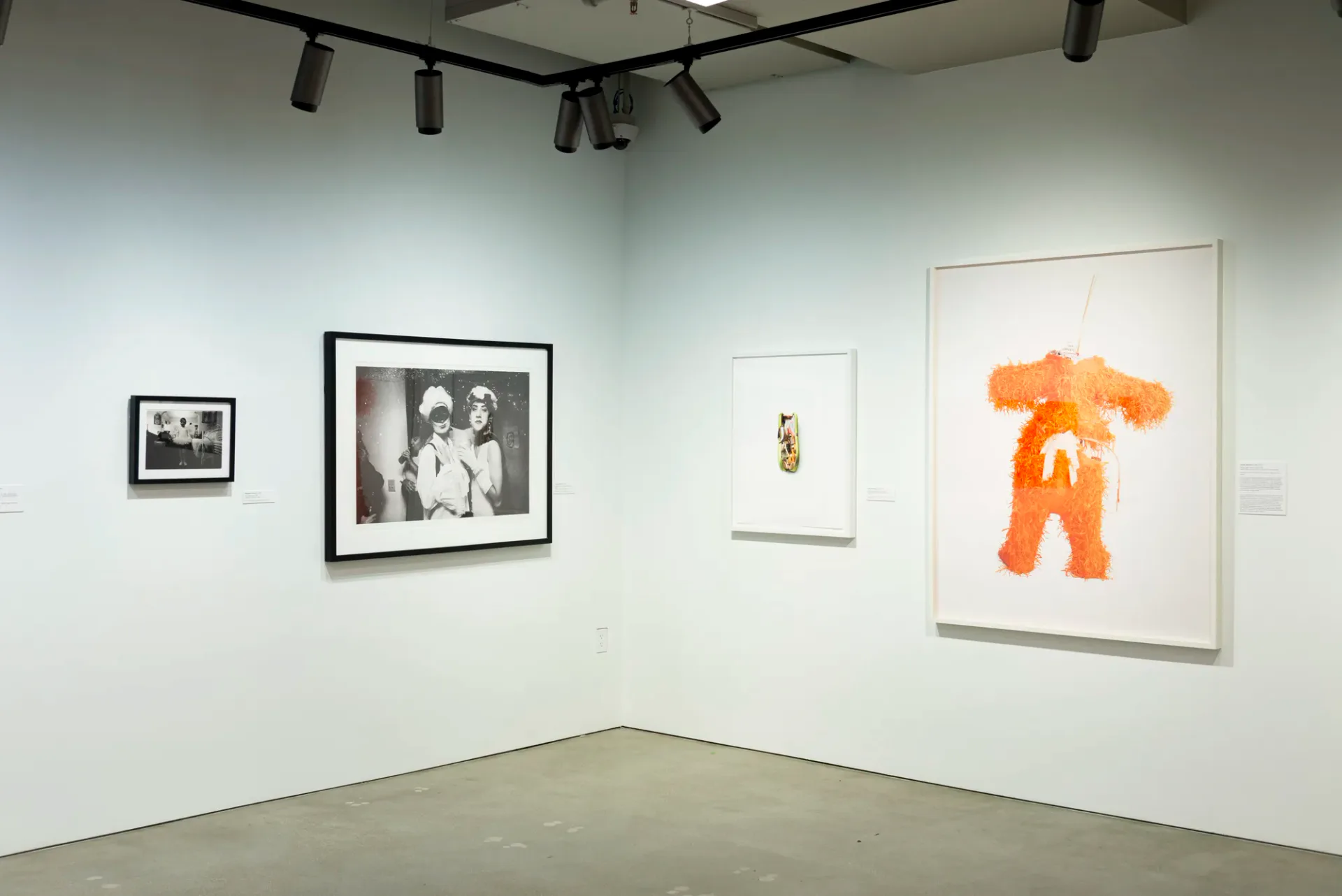 Corner gallery view with black-and-white photographs and a bright orange abstract figure drawing on white wall.