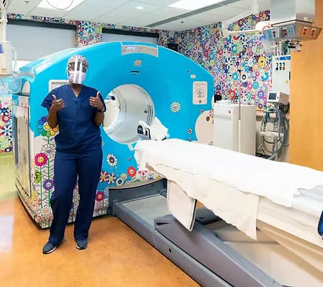 Interior of RX Art gallery showing a colorful wrapped MRI scanner machine decorated with sticker art, medical staff member standing beside it, medical equipment and treatment room visible.