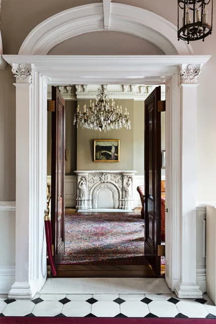 Grand white neoclassical archway with Corinthian capitals opening onto formal room with fireplace, chandelier, and patterned rug at Salmagundi Club.