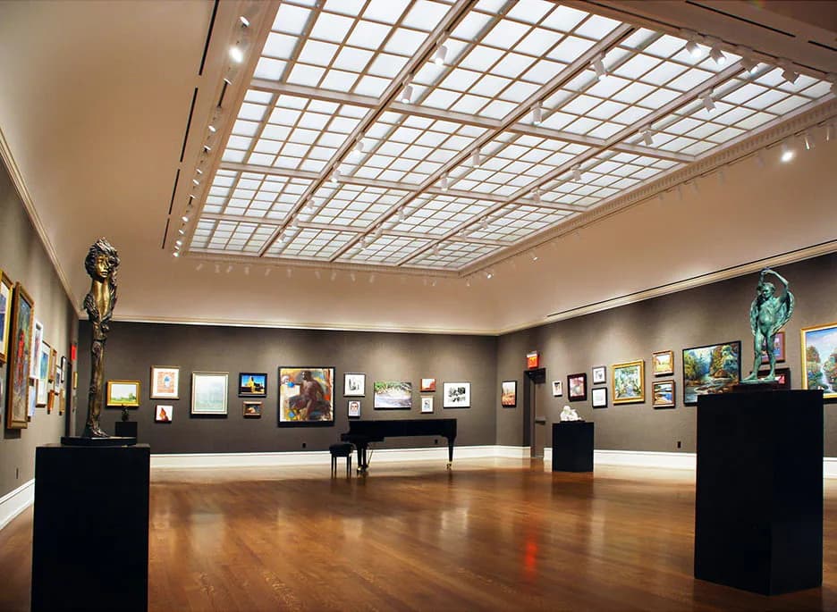 Modern art gallery interior with grid skylight ceiling, dark walls displaying framed artworks, polished wooden floor, and black pedestals.