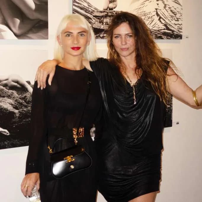 Two women posing together indoors against a backdrop of black and white artwork; one wears blonde hair and red lips, the other has dark hair.