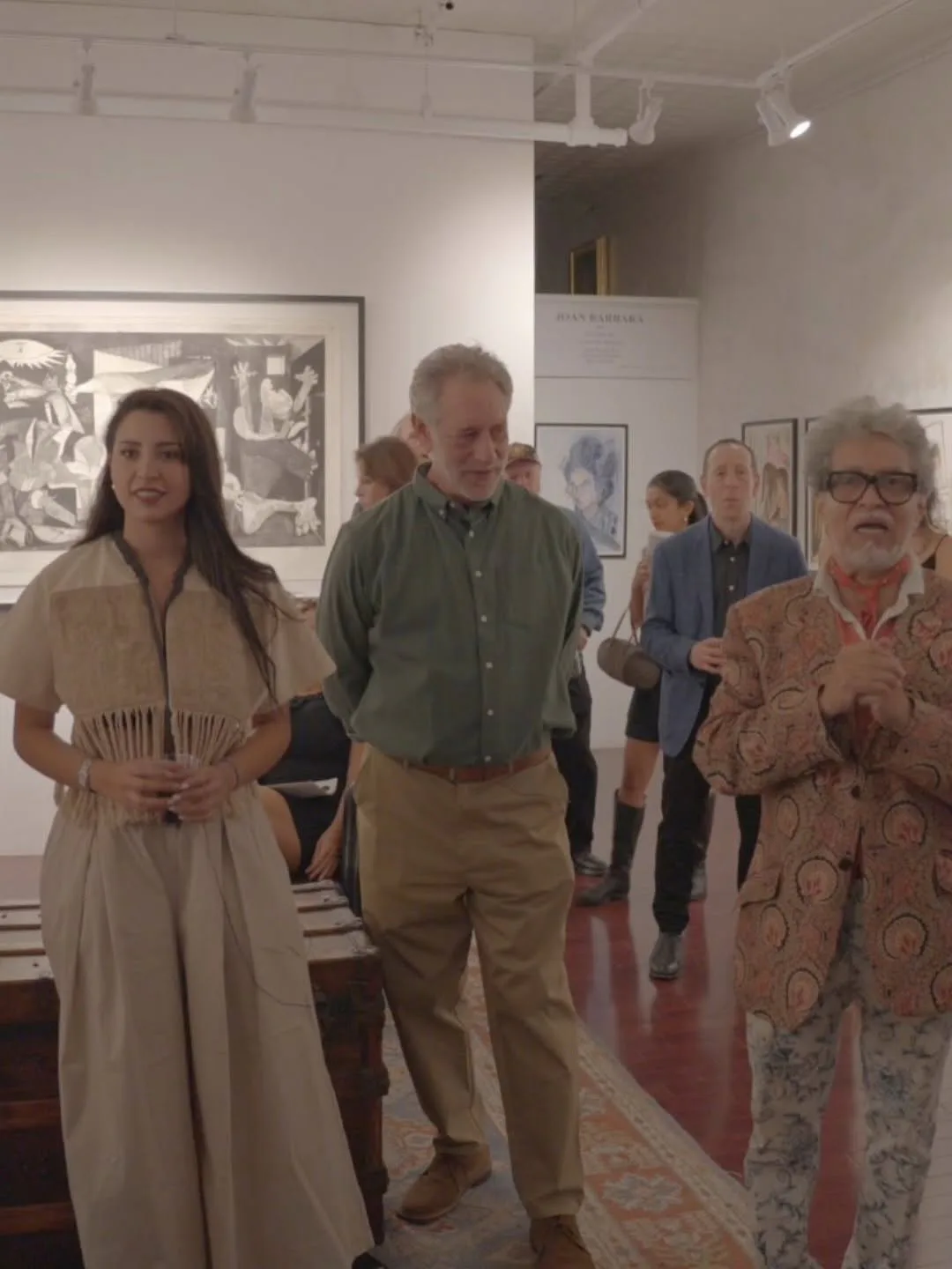 Gallery interior with four people standing among framed artworks on white walls and a patterned floor, viewing an exhibition.