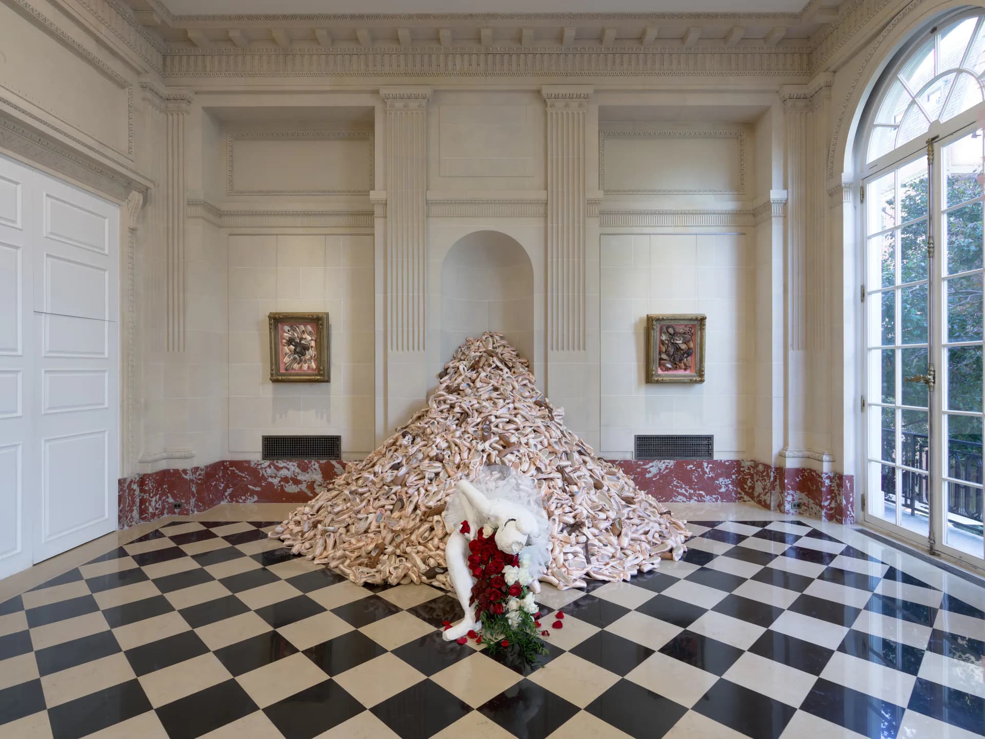 Installation in a neoclassical room with black-and-white checkerboard floor: a large conical mound of pale wood chips or shavings with a single red flower at its base.