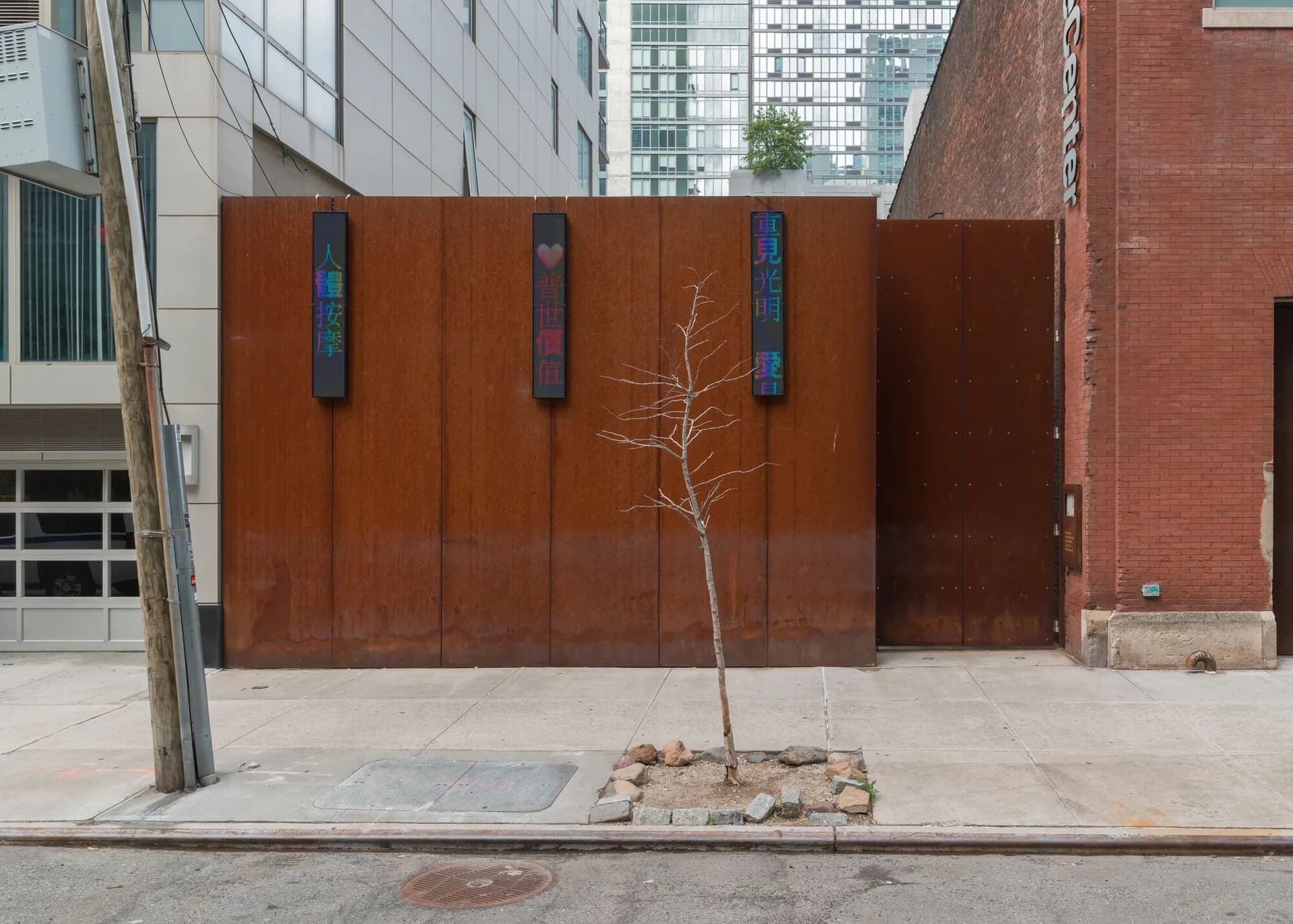 Outdoor plaza at Sculpture Center featuring rust-colored corrugated metal wall with three vertical openings and a young bare tree.