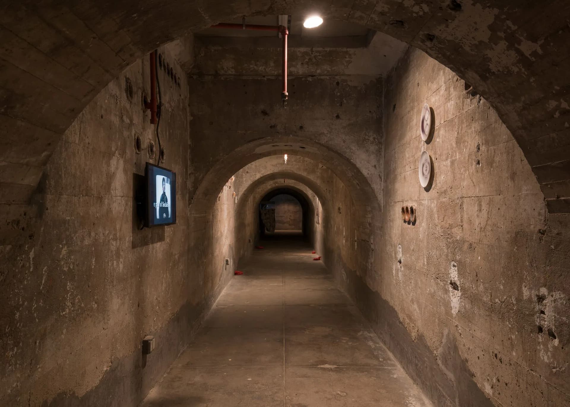 Arched tunnel passageway with concrete walls and archways receding into distance, single overhead light, small blue monitor visible.