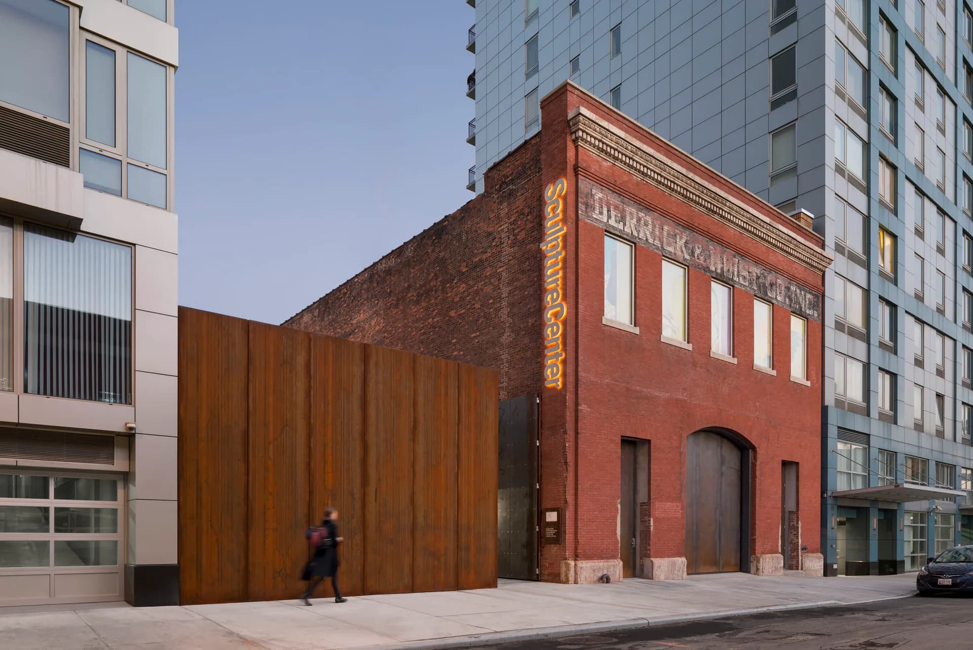 Exterior of SculptureCenter in Long Island City showing a red brick building with arched windows adjacent to a large weathered steel panel structure on a concrete plaza.