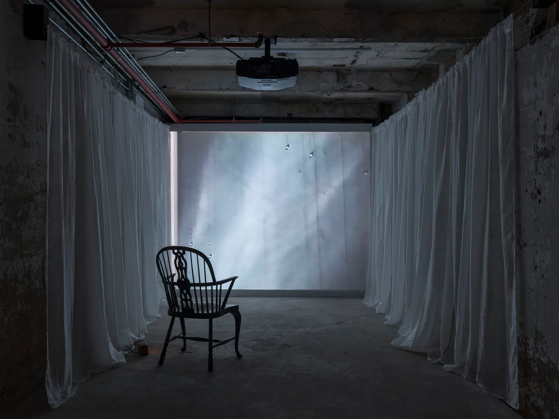 Installation artwork: sparse concrete bunker-like space with sheer white curtains framing a glowing window, single black Windsor chair positioned centrally, creating contemplative atmosphere.
