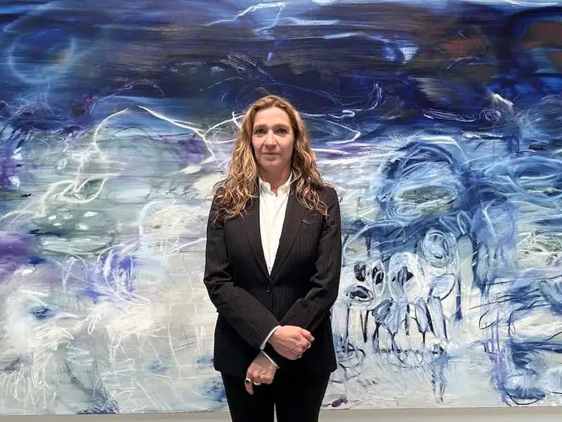 Woman in dark blazer and white shirt standing in front of an abstract blue and white painted mural or wall installation in a gallery space.