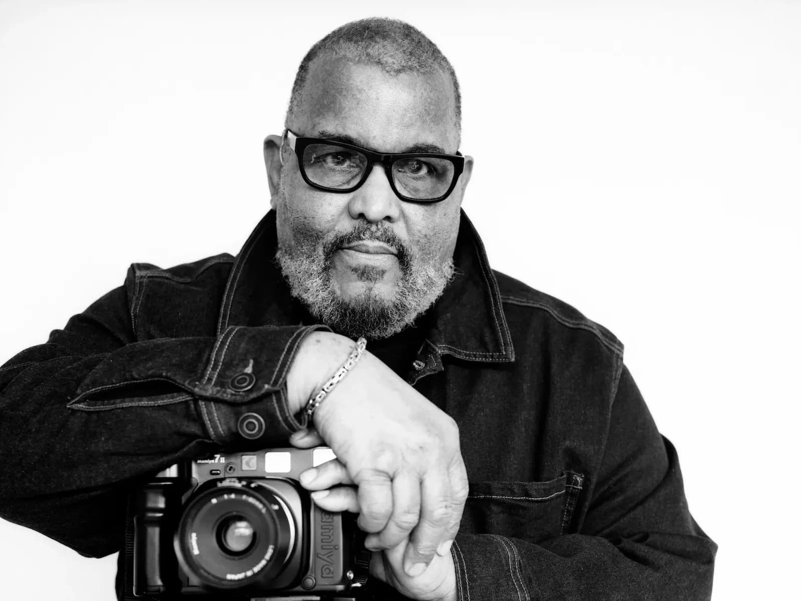 Black and white portrait of a man wearing glasses and dark jacket, holding a vintage camera, against a neutral background.