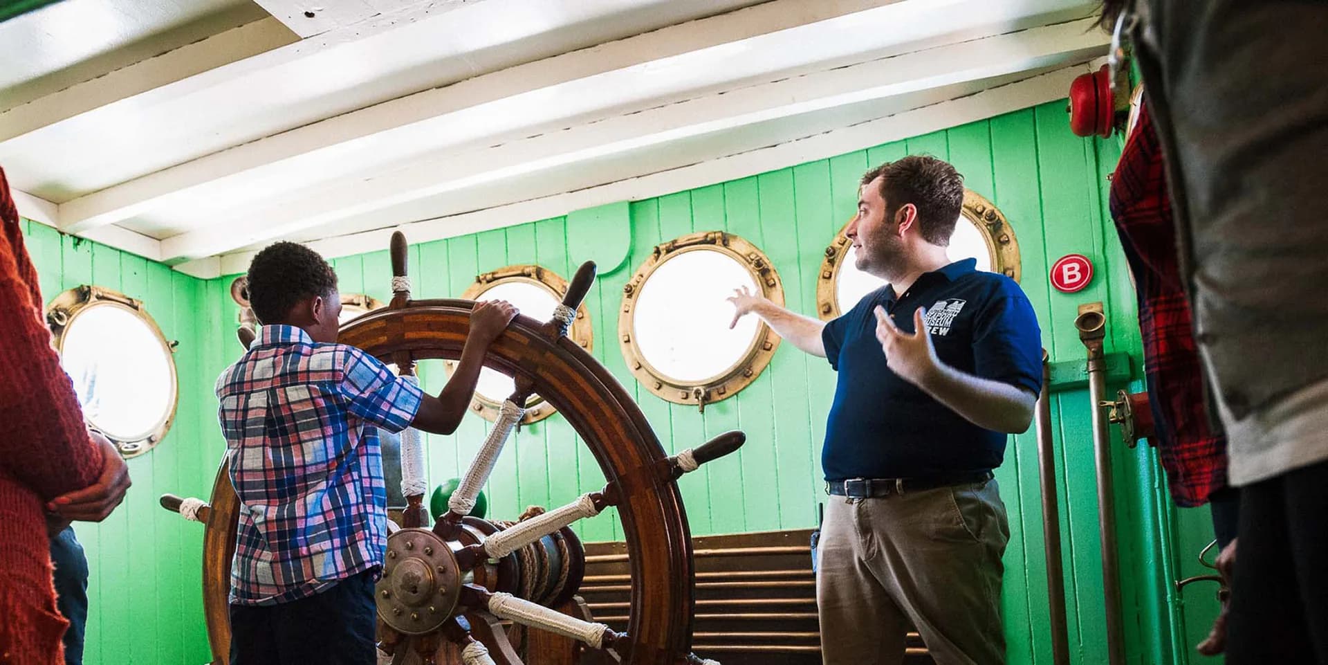 Interior of a museum gallery with green walls, visitors interacting with a large wooden ship's wheel and brass portholes.