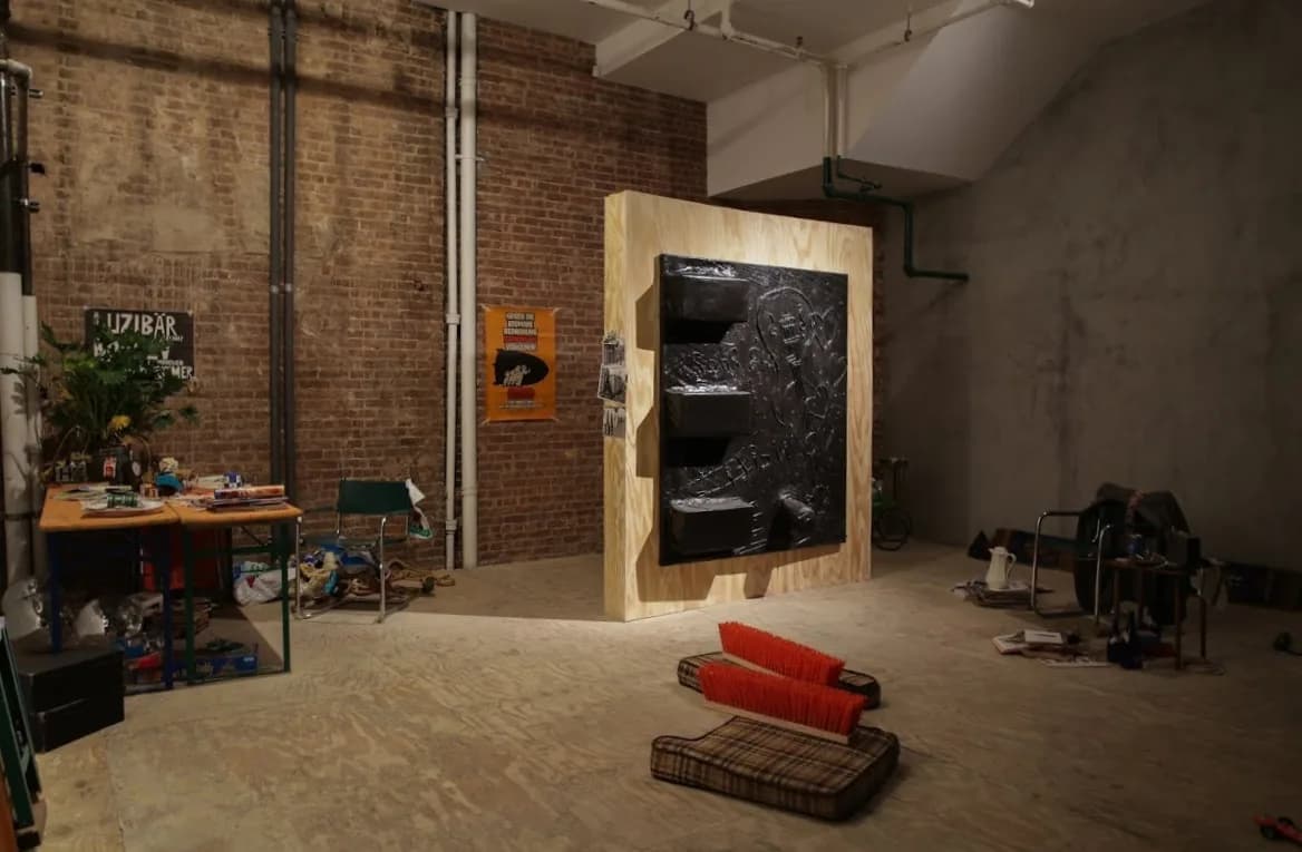 Gallery interior with exposed brick wall, wooden sculpture installation, worktables, and scattered objects on the floor