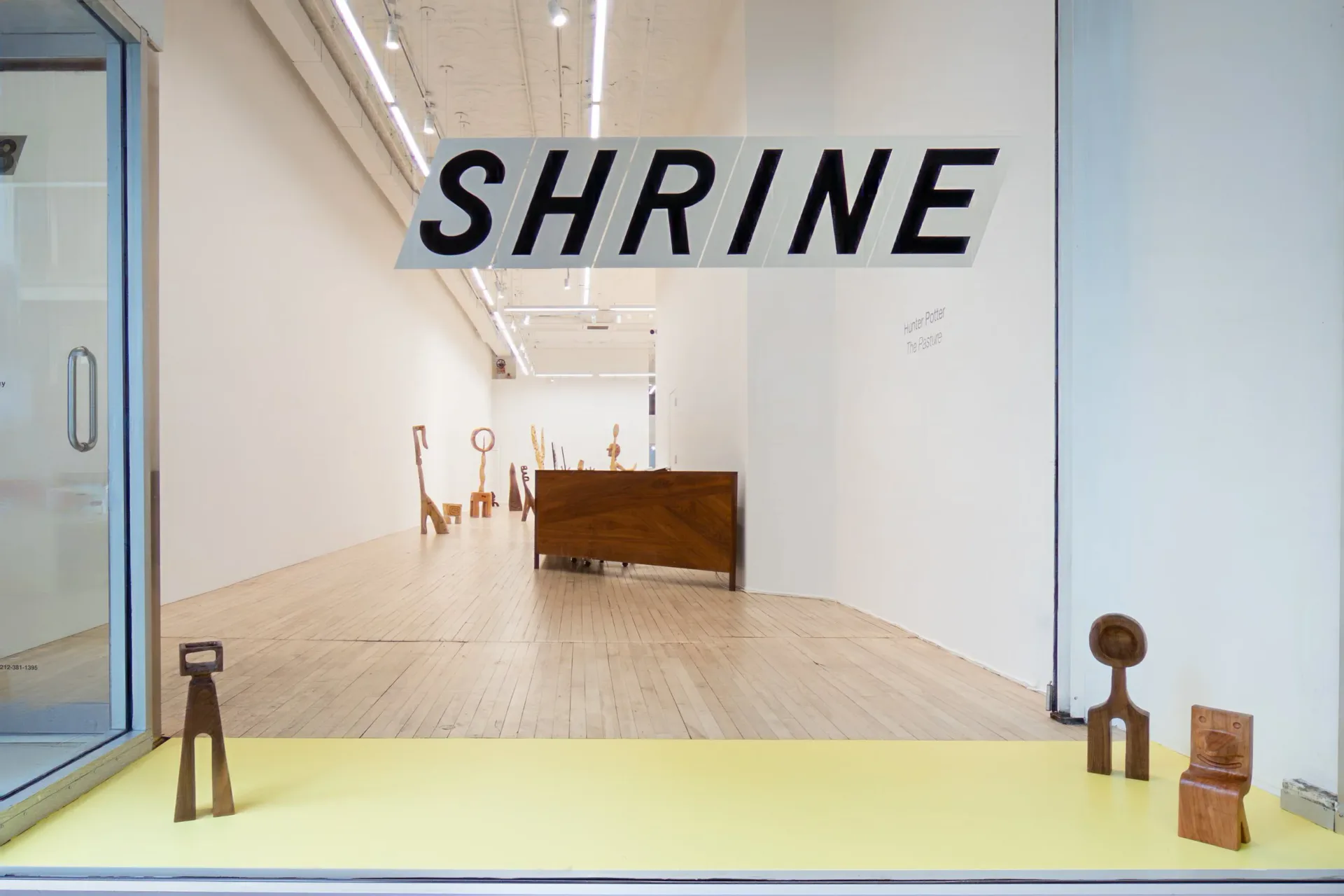 Gallery interior with white walls, wood floor, hanging SHRINE sign, and small wooden sculptures on a yellow display shelf