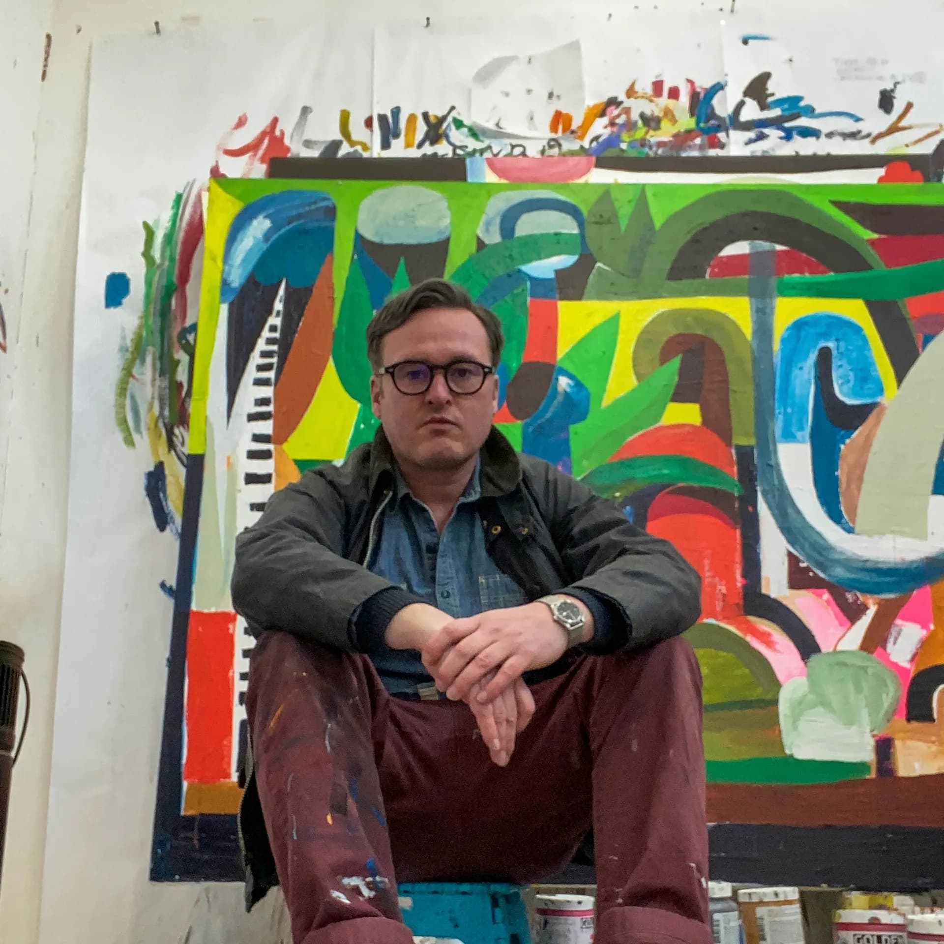 Male artist wearing glasses and dark jacket seated before colorful abstract mixed-media artwork with geometric shapes and text elements.