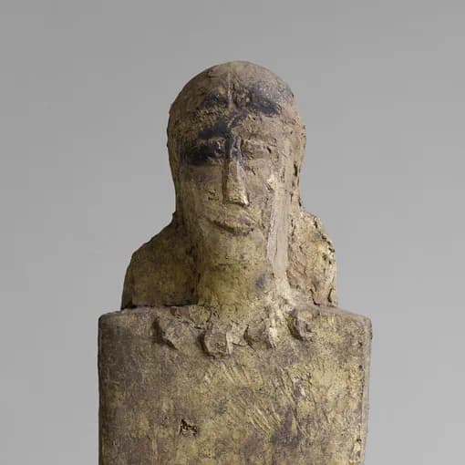 Stone or concrete sculptural bust depicting a stylized human head with simplified features mounted on a rectangular base.