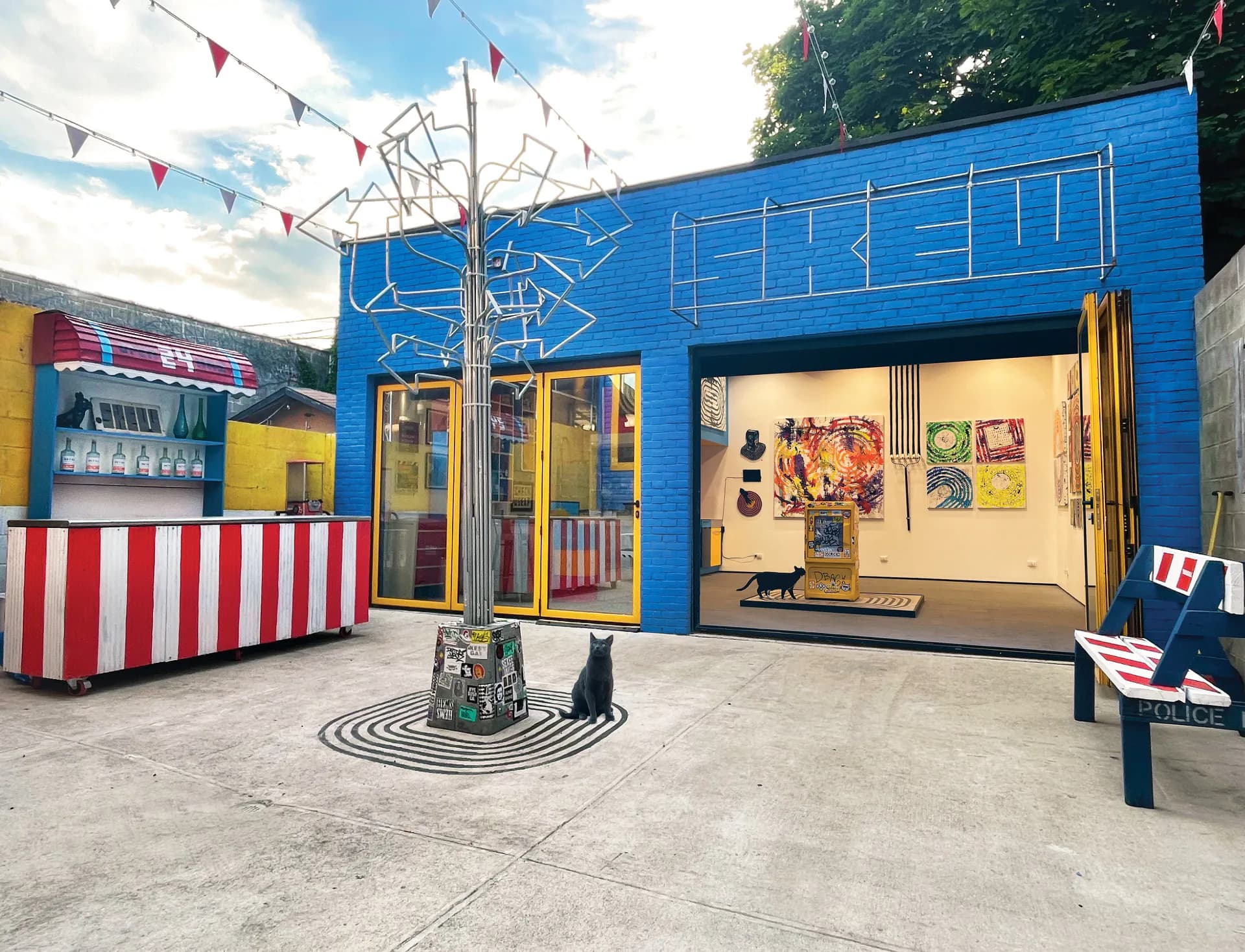 Exterior of SKEWVILLE gallery storefront with bright blue facade, yellow doors, red and white striped booth, hanging sneakers, and artwork visible inside.